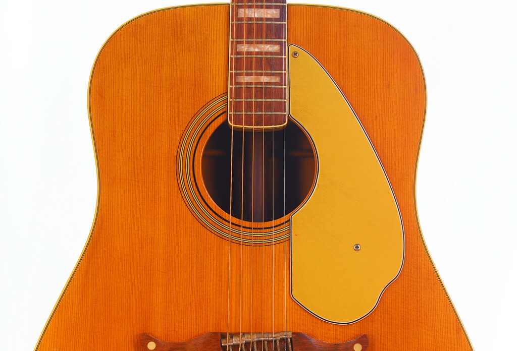 Close-Up Of A Fender Wildwood 1967 Acoustic Guitar, Featuring A Natural Wood Finish, Teardrop-Shaped Yellow Pickguard, Visible Strings, And Soundhole Set Against A Plain White Background.