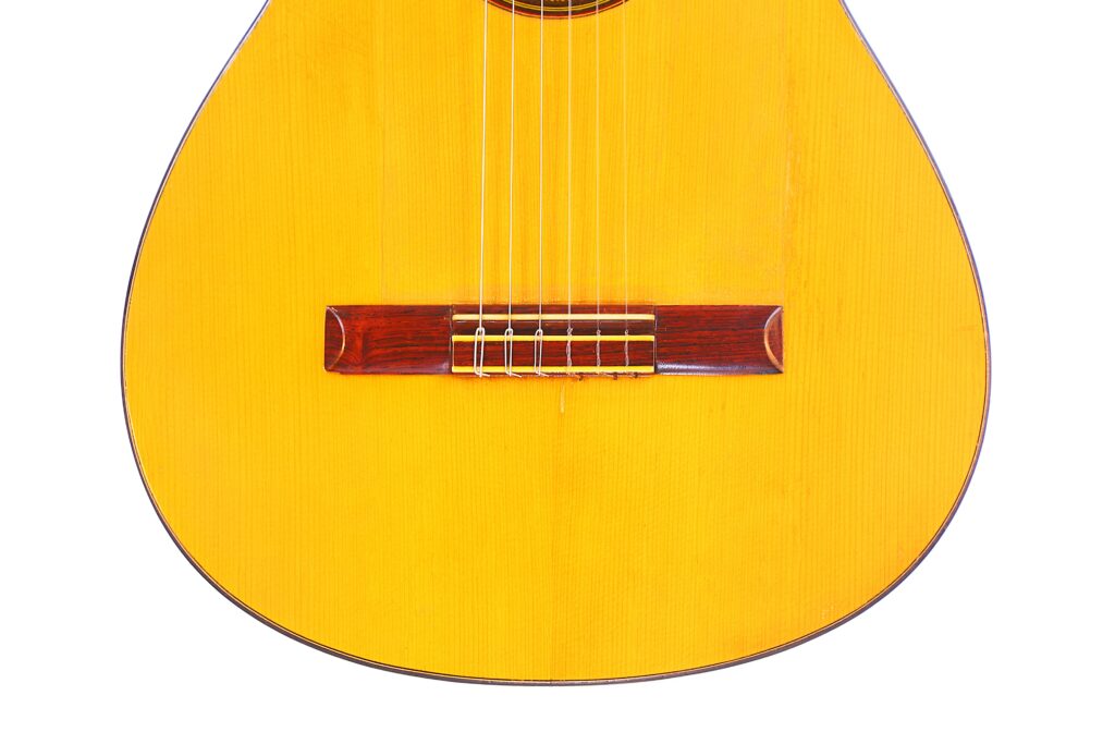 Close-Up Of The Lower Half Of A Conde Hermanos 1971 Flamenco Guitar, Featuring Its Light Wood Body, Six Strings, And Dark Wooden Bridge Against A White Background.