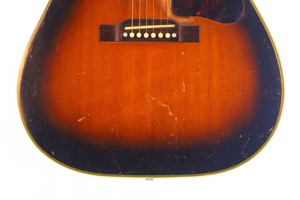 Close-Up Of A Gibson Southern Jumbo 1955 Acoustic Guitar Body, Showing Its Sunburst Dark Brown To Amber Finish, Bridge, Strings, And Visible Scratches That Highlight Its Vintage Character.