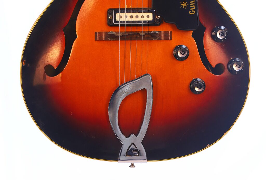 Close-Up Of The Guild T-100D Slim Jim 1967 Electric Guitar Featuring Two F-Shaped Sound Holes, Four Black Knobs, A Pickup, And A Distinctive Silver Tailpiece On A White Background.