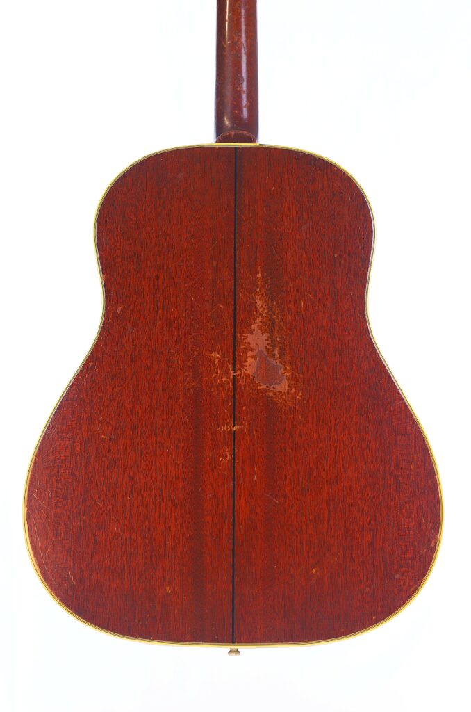 The Back Of The Gibson Southern Jumbo 1955 Acoustic Guitar Features A Reddish-Brown Wood Finish With Visible Grain, Some Scratches, And A Thin Dark Center Seam, Displayed Against A Plain White Background.