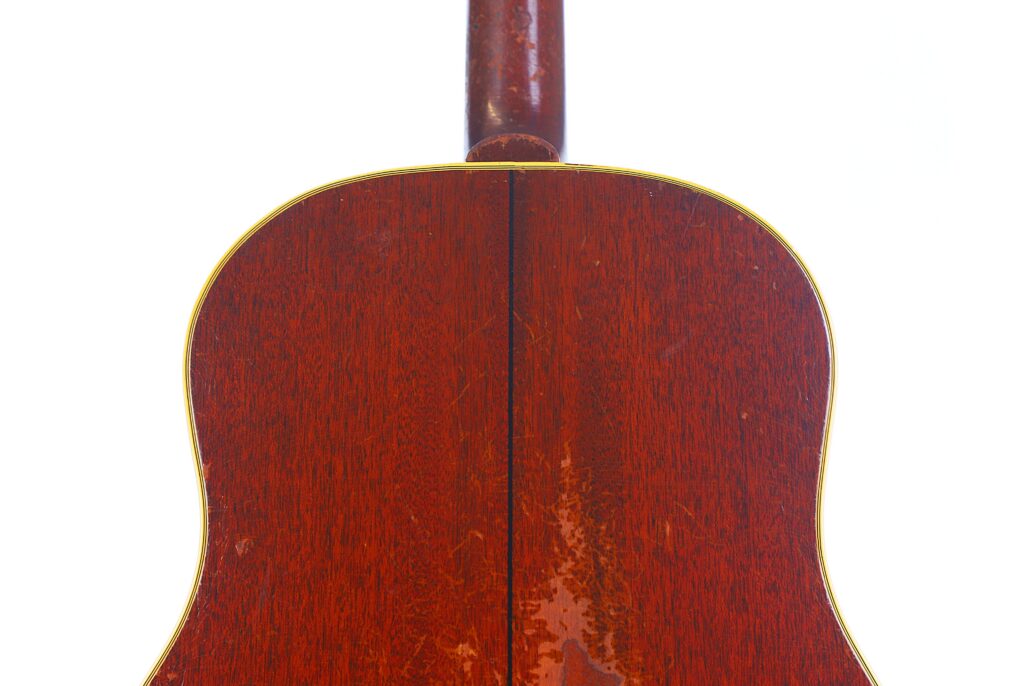 Close-Up Of The Back Of A Gibson Southern Jumbo 1955 Acoustic Guitar, Featuring Its Reddish Wood Finish With A Yellow Outline And Center Seam, Plus The Neck Visible At The Top Against A White Background.