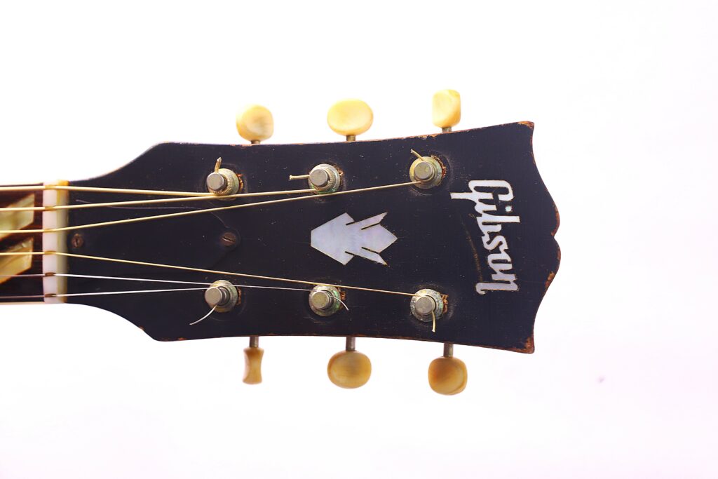 Close-Up Of The Black Headstock With Cream Tuners, Strings, And White Gibson Logo On The Gibson Southern Jumbo 1955 Acoustic Guitar, Set Against A White Background.