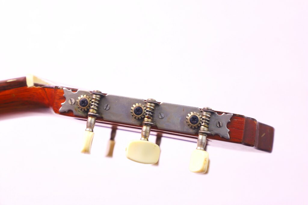 Close-Up View Of The Tuning Pegs And Gears On The Headstock Of A Conde Hermanos 1971 Flamenco Guitar Against A Light Pink Background.