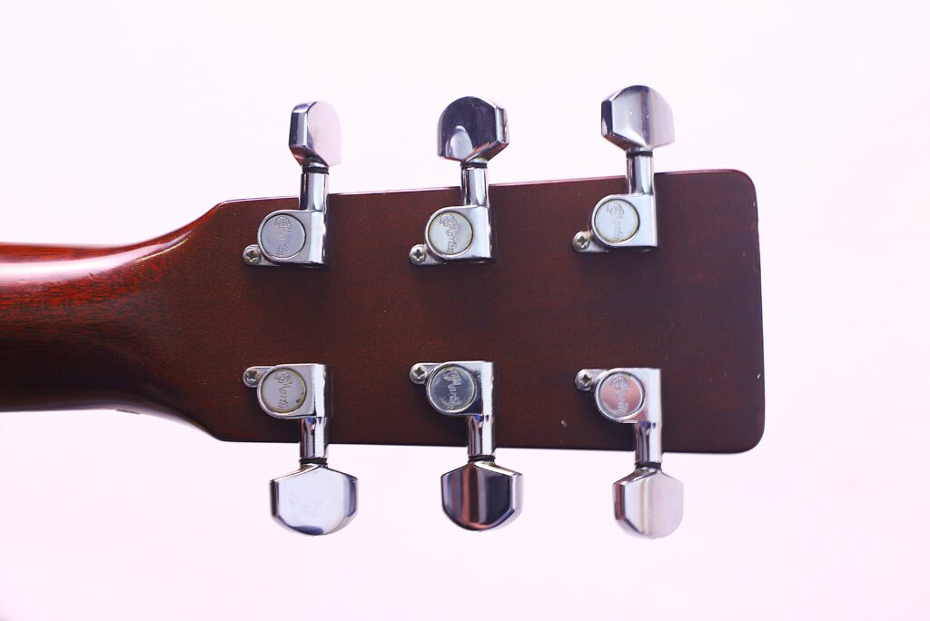 Close-Up Of The Back Of A Martin Hd-35 1979 Acoustic Guitar Headstock, Displaying Six Chrome Tuning Pegs On Its Wooden Neck, Set Against A Plain Light Pink Background.