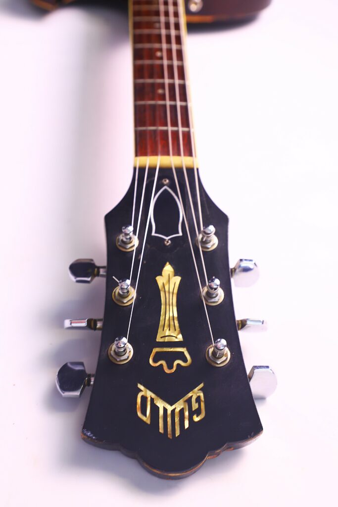 Close-Up Of The Guild T-100D Slim Jim 1967 Electric Guitar Headstock, Showing Six Tuning Pegs And Gold Details On A Dark Background, Photographed Against A White Surface.