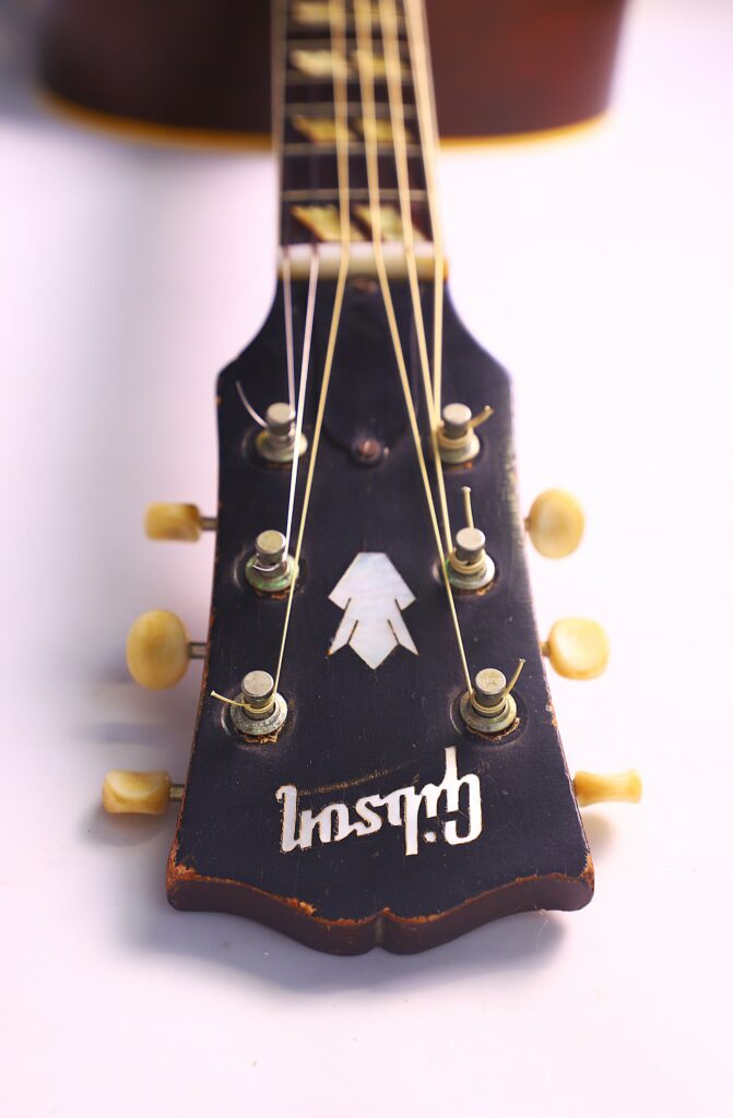 Close-Up Of The Gibson Southern Jumbo 1955 Acoustic Guitar Headstock With Tuning Pegs, Strings, And Gibson Logo, Focusing On The Top Of The Neck Against A Light Background.