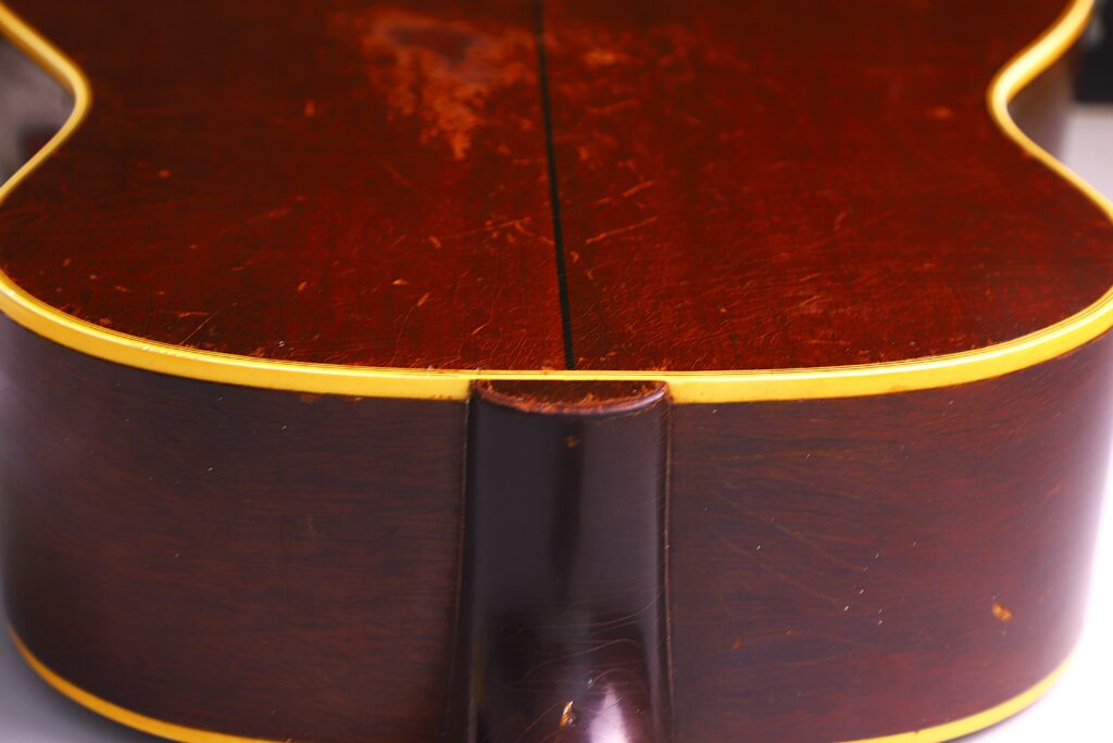 Close-Up Of The Back Of A Gibson Southern Jumbo 1955 Acoustic Guitar, Featuring A Dark Wood Finish With A Central Seam, Yellow Edge Binding, And A Partial View Of The Neck Joint At The Bottom.