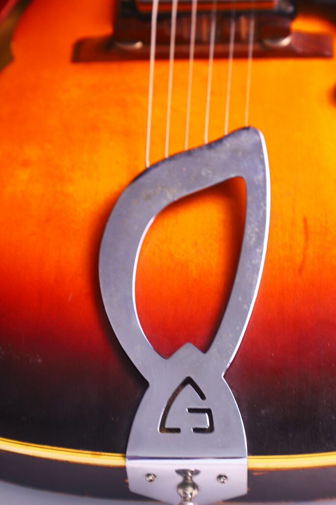 Close-Up Of The Guild T-100D Slim Jim 1967 Electric Guitar’s Sunburst Body, Showcasing Its Unique Silver Metal Tailpiece And Strings, With Emphasis On The Distinctive Lower Edge Design.