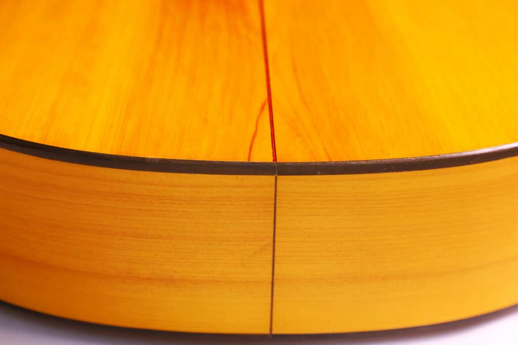 Close-Up Of The Bottom Edge Of A Conde Hermanos 1971 Flamenco Guitar, Highlighting Its Smooth, Glossy Wooden Surface And The Seam Where Two Wood Pieces Meet, With Light Reflecting Off The Polished Finish.