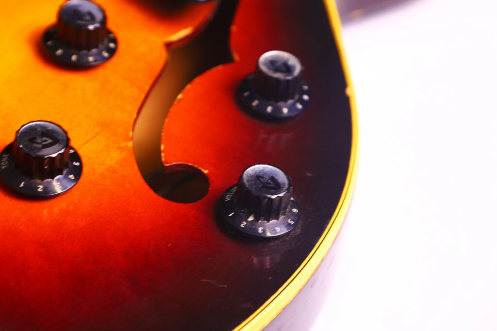 Close-Up Of The Guild T-100D Slim Jim 1967 Electric Guitar Body, Featuring Four Black Control Knobs And Part Of An F-Hole On A Glossy Orange-Brown Sunburst Finish With A Yellow-Edged Border.