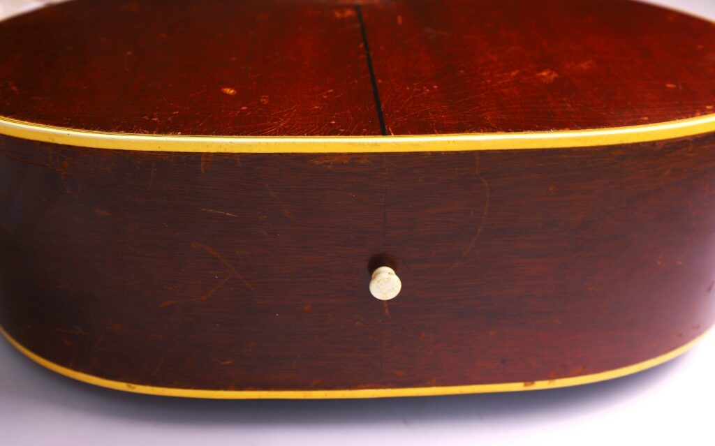 Close-Up Of The Bottom Side Of A Gibson Southern Jumbo 1955 Acoustic Guitar, Highlighting Its Metal Strap Button, Glossy Reddish-Brown Finish With Scratches, And Distinctive Yellow Trim.