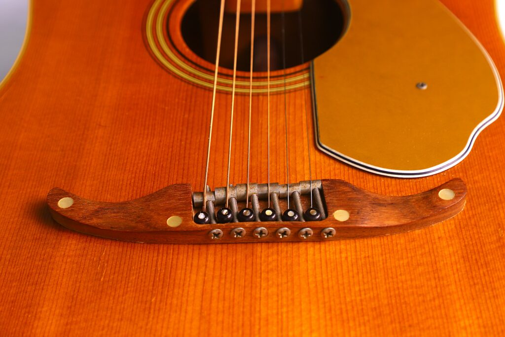 Close-Up Of A Fender Wildwood 1967 Acoustic Guitar’s Wooden Bridge, Strings, And Part Of The Golden Pickguard On Its Polished Orange-Brown Body.