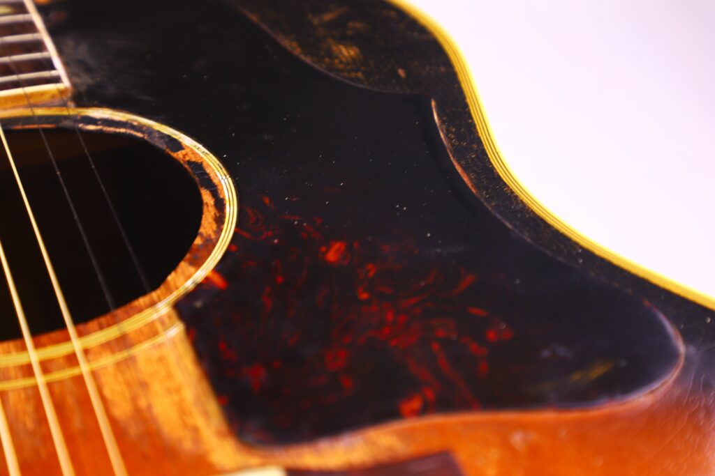Close-Up Of The Gibson Southern Jumbo 1955 Acoustic Guitar’s Body, Featuring Worn Wood, A Dark Pickguard With Red Patterns, Gold Strings, And Part Of The Sound Hole, Highlighting Its Unique Texture And Vintage Character.