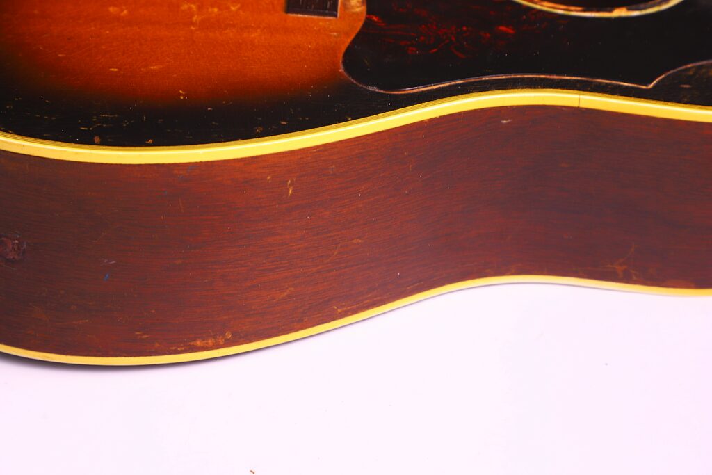 Close-Up Of The Gibson Southern Jumbo 1955 Acoustic Guitar, Highlighting Its Sunburst Top, Rich Brown Wooden Body, And Yellow Binding, Set Against A Plain White Background.
