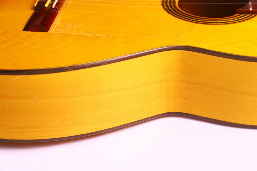 Close-Up Of The Side And Body Of A Conde Hermanos 1971 Flamenco Guitar In Yellow, Showing The Wood Grain, Part Of The Sound Hole, And Bridge, Against A White Background.