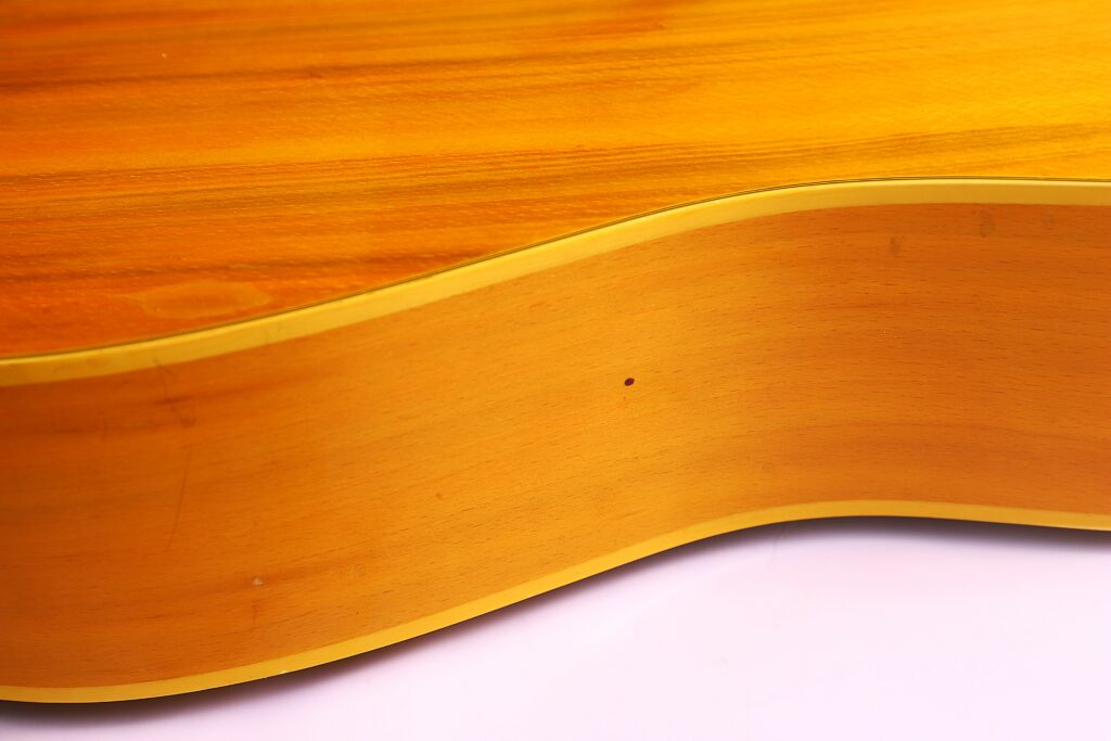 Close-Up Of The Side Of A Fender Wildwood 1967 Acoustic Guitar, Highlighting Its Smooth Light Brown Wood Finish, Cream Binding, Subtle Wood Grain Texture, And A Small Dark Dot On The Side.