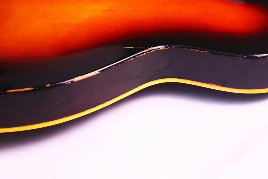Close-Up Of The Side Of A Guild T-100D Slim Jim 1967 Electric Guitar, Showing Worn Edges And Paint Chips Along Its Smooth Curves Against A White Background.