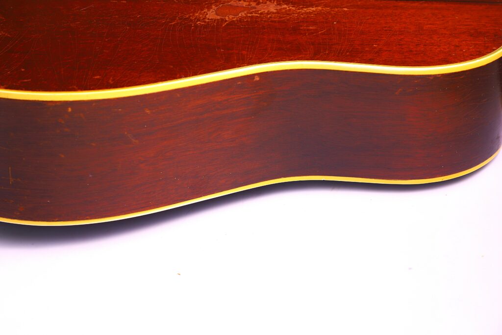 Close-Up Of The Gibson Southern Jumbo 1955 Acoustic Guitar’s Side, Featuring A Glossy Brown Finish And Yellow Trim, Set Against A White Background.