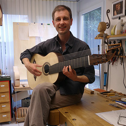 A Smiling Man Sits On A Workbench In A Workshop, Holding A Large, Multi-String Classical Guitar. Various Tools And Woodworking Materials Are Visible Around Him, And Natural Light Comes In Through A Window.