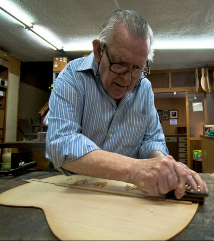 Manuel Reyes Wearing Glasses And A Striped Shirt Shapes A Piece Of Wood With A Hand Tool In A Workshop, Possibly Crafting A Musical Instrument Like A Guitar.