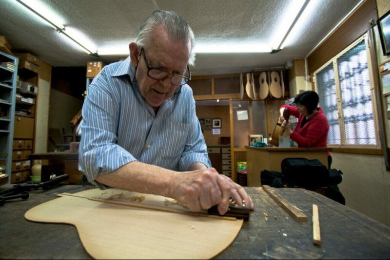 Manuel Reyes Shapes A Wooden Guitar Top In A Workshop, Focused On His Craft. In The Background, A Woman Uses A Tool At Another Workbench. Shelves With Guitar Parts And Tools Fill The Room.