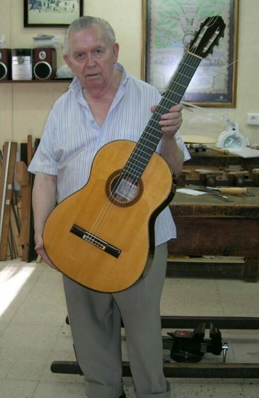An Elderly Man With Gray Hair, Wearing A Light Striped Shirt And Gray Pants, Stands Indoors Holding A Classical Acoustic Guitar Upright In Front Of Him. Workshop Tools And Wooden Workbenches Are Visible In The Background.