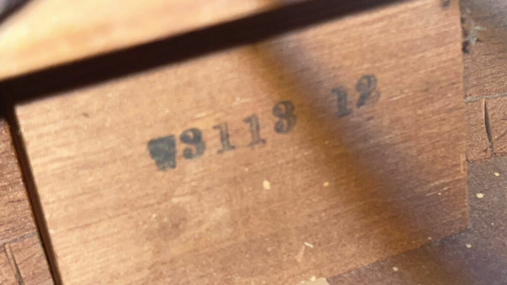 Close-Up Of The Wooden Surface Of A Gibson Southern Jumbo 1955 Acoustic Guitar, Showing Black-Ink Stamp “3113 12” Partially Hidden By Shadow And Small Wood Particles Visible.