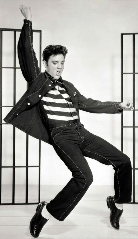 A Man Dressed In A Black Jacket And Striped Shirt Dances Energetically In Front Of Metal Bars, With One Arm Raised And One Bent, Mid-Jump, Evoking A Classic Rock And Roll Performance.