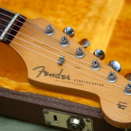 Close-Up Of A Fender Stratocaster Guitar Headstock With Tuning Pegs, Logo, And Text Visible, Resting Inside A Yellow Plush-Lined Brown Guitar Case.