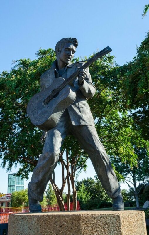 A Bronze Statue Of A Man Playing Guitar Stands Outdoors On A Pedestal, Surrounded By Green Trees And Clear Blue Sky. The Man Is Posed In A Dynamic, Wide-Legged Stance, Wearing A Suit.