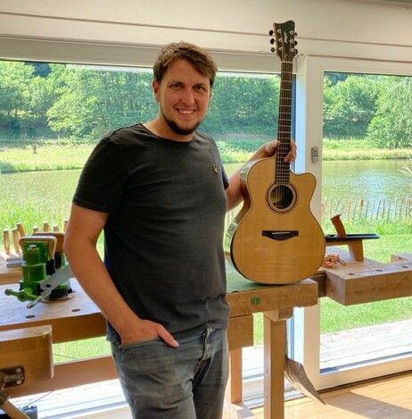 A Smiling Man Stands Indoors By A Workbench, Holding An Acoustic Guitar Upright, Ready For Guitar Repair. Large Windows Behind Him Reveal A Scenic View Of Greenery And A Pond Outside.
