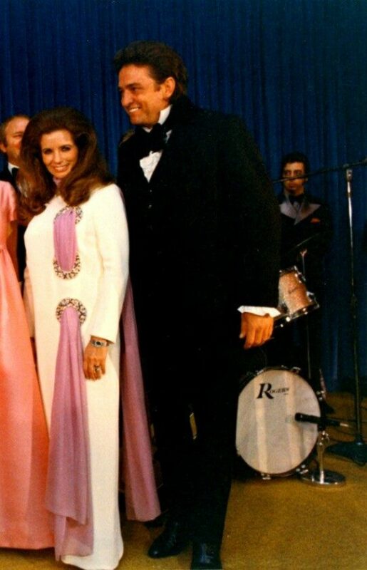 A Woman In A White And Purple Gown Stands Next To A Man In A Black Tuxedo, Both Smiling, In Front Of A Blue Curtain With A Drummer And Band Visible In The Background.