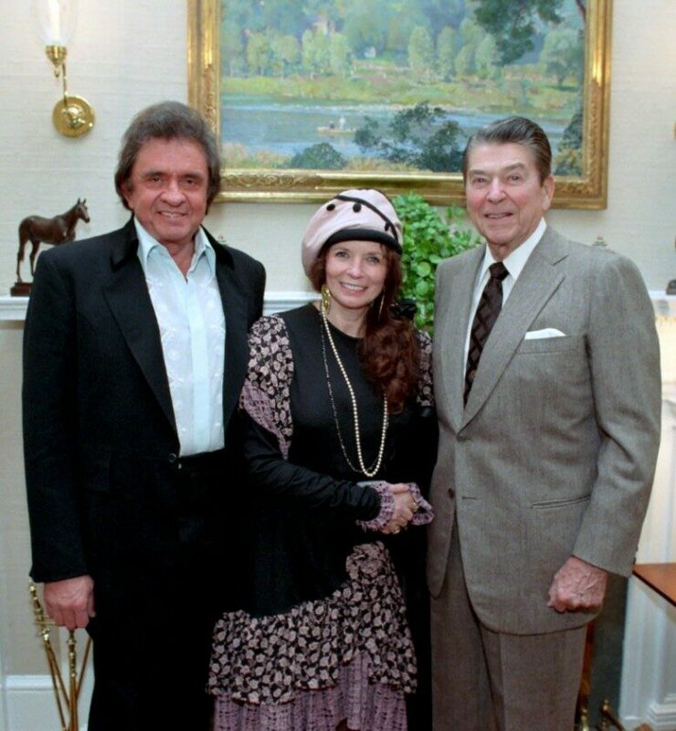Three Adults Stand Together In A Formal Room. The Man On The Left Wears A Dark Suit, The Woman In The Center Wears A Pink And Black Dress With A Matching Hat, And The Man On The Right Wears A Grey Suit And Striped Tie. A Painting Hangs Behind Them.