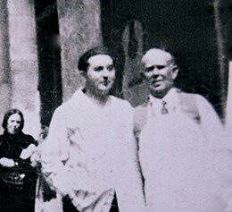 A Vintage Black-And-White Photo Shows Two Men Standing Close Together, Both Wearing Light-Colored Clothing. In The Background, A Woman In Dark Clothes Walks Past Large Columns. The Image Has A Blurry, Old-Fashioned Appearance, Reminiscent Of Domingo Esteso’s Era.