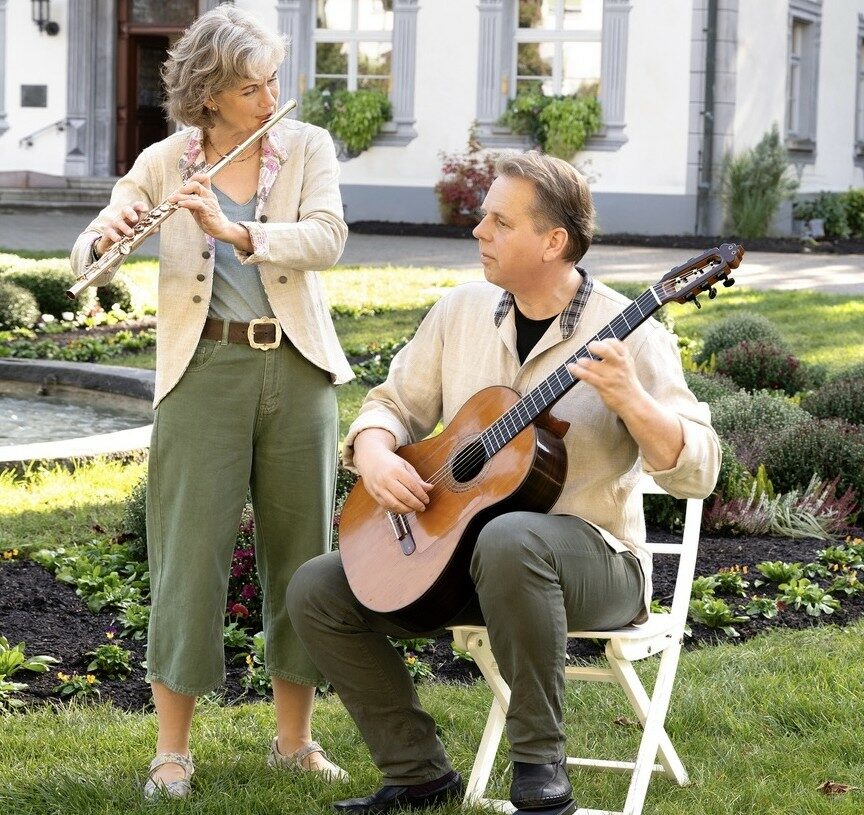 A Woman Plays The Flute While Standing Next To A Man Sitting On A White Chair, Playing Guitar In A Garden With A Fountain And Large Building—Offering Their Musical Service. Both Are Dressed In Light-Colored Clothing.