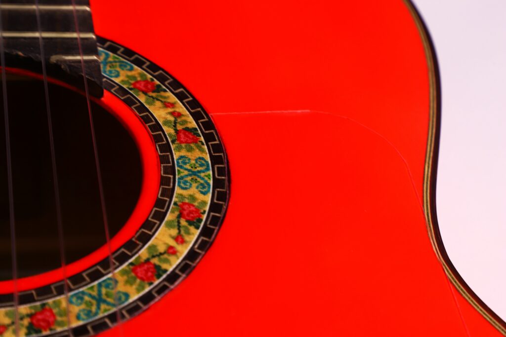 Close-Up Of A Pedro Fernández Luque 2021 Blanca Flamenco Guitar In Bright Red, Featuring Part Of The Sound Hole With A Colorful Floral Pattern, Visible Strings, And A Glossy Body.