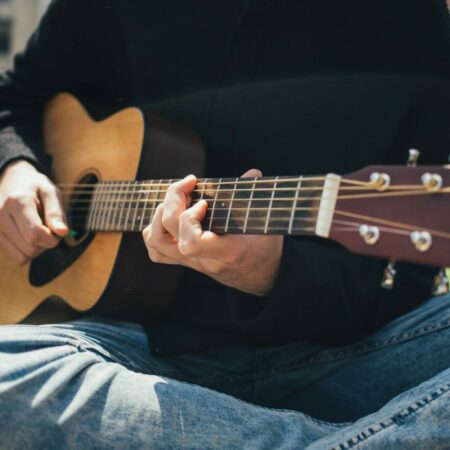 Person Sitting Cross-Legged Outdoors, Wearing Jeans And A Black Sweater, Playing An Acoustic Guitar. The Focus Is On Their Hands And The Guitar, With A Blurred Urban Background.