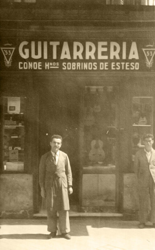 Two men stand outside a guitar shop with a sign reading “GUITARRERIA CONDE HNOS SOBRINOS DE ESTESO.” The shopfront features display windows with guitars and other items. The image is sepia-toned and vintage.