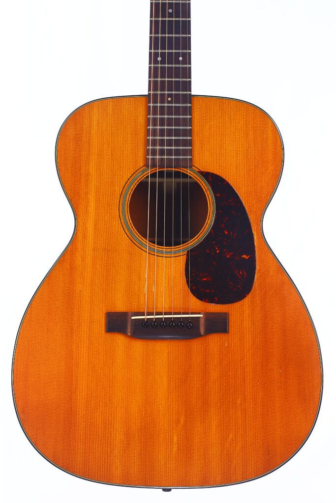 Die Martin 000-18 1958 Akustik Gitarre Verfügt Über Ein Naturholz-Finish, Sechs Saiten, Ein Schwarzes Schlagbrett Und Dekorative Ringe Um Das Schallloch. Sie Wird Von Vorne Vor Einem Weißen Hintergrund Gezeigt.