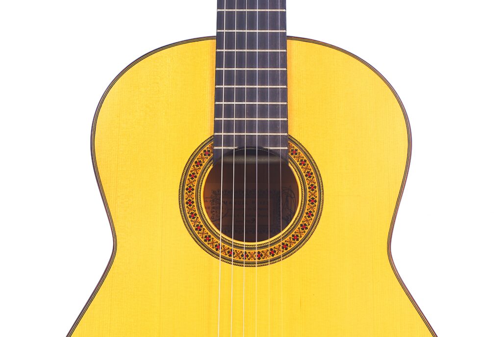 Nahaufnahme Des Korpus Und Des Griffbretts Einer Manuel Reyes Flamenco-Gitarre Aus Dem Jahr 1991, Wobei Das Gelbe Holzfinish, Die Saiten Und Die Dekorative Rosette Um Das Schallloch Vor Weißem Hintergrund Hervorgehoben Werden.