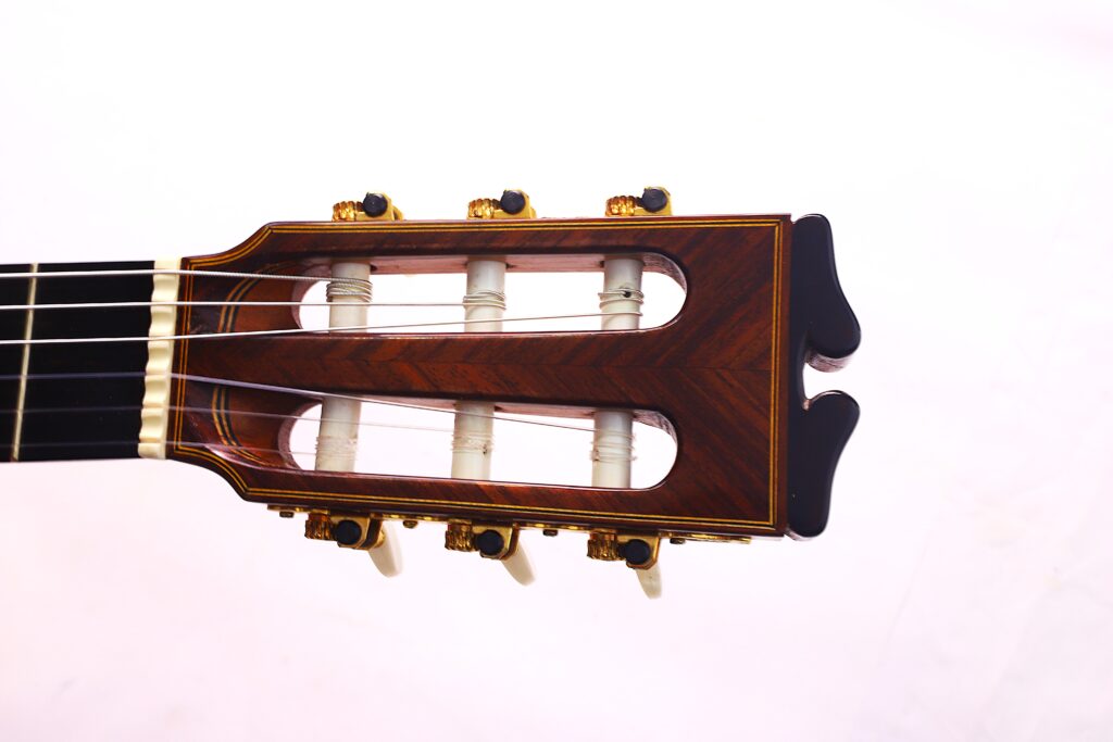 Nahaufnahme Der Kopfplatte Der Luis Sanchez 2011 Negra Flamenco Gitarre Mit Dunkler Holzoberfläche, Goldenen Stimmwirbeln Und Weißen Stiften Vor Einem Weißen Hintergrund. Die Gitarrensaiten Verlaufen Entlang Der Kopfplatte.