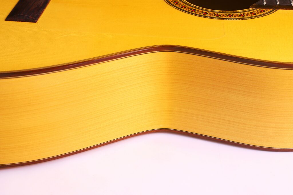 Nahaufnahme Der Seite Und Oberfläche Einer Manuel Reyes 1991 Flamenco-Gitarre, Die Die Helle Holzmaserung, Den Gebogenen Korpus Und Einen Teil Des Schalllochs Mit Dekorativer Einlage Hervorhebt.