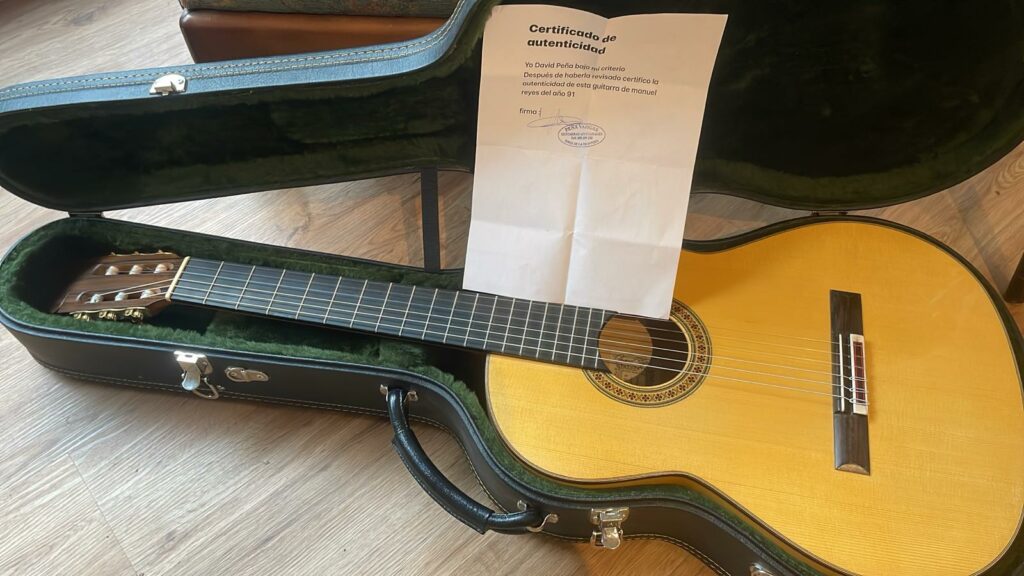 Eine Manuel Reyes 1991 Flamenco-Gitarre Ruht In Einem Offenen, Schwarzen Hartschalenkoffer, Der Mit Grünem Samt Ausgekleidet Ist, Auf Dem Ein Echtheitszertifikat Ruht. Das Gehäuse Steht Auf Einem Holzboden.