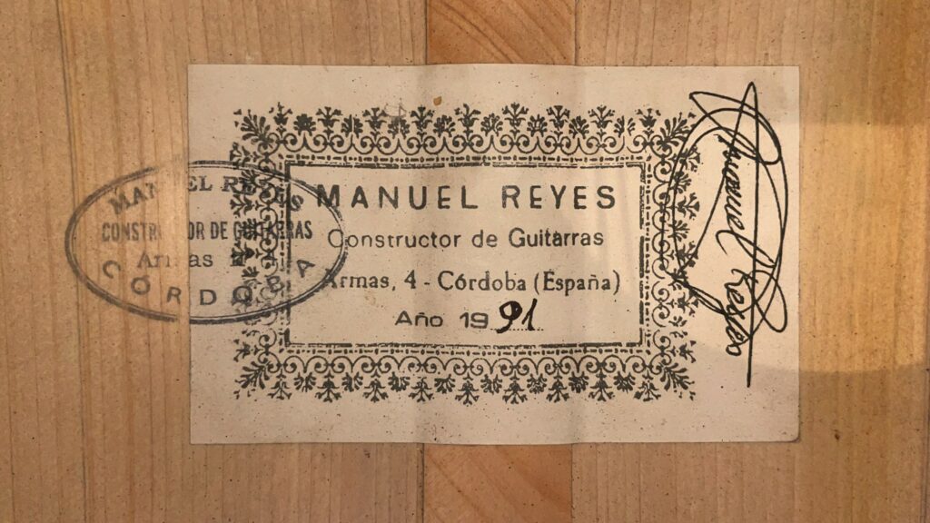 Die Manuel Reyes 1991 Flamenco-Gitarre Verfügt Über Ein Vintage-Etikett Mit Der Aufschrift: &Quot;Manuel Reyes, Constructor De Guitarras, Armas, 4 - Córdoba (España), Año 1991&Quot; Mit Einer Dekorativen Umrandung, Einem Kreisförmigen Stempel Und Einer Signatur Auf Einer Holzoberfläche.