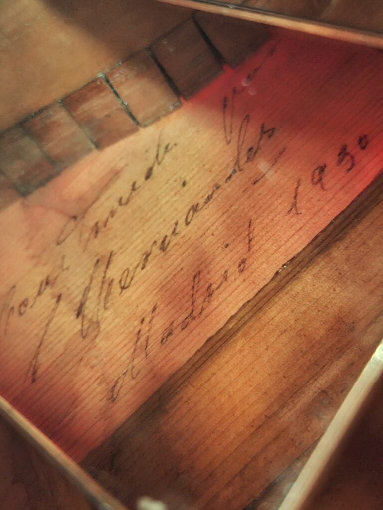Nahaufnahme eines handgeschriebenen Etiketts im Inneren eines hölzernen Musikinstruments mit der Aufschrift „Jose Garcia Hernandez, Madrid 1930“. Das Bild zeigt Details des Instrumenteninneren und charakteristische Merkmale, die an die für die Flamenco-Gitarre von Santos Hernandez aus dem Jahr 1930 typische Handwerkskunst erinnern. Nahaufnahme eines handgeschriebenen Etiketts im Inneren eines hölzernen Musikinstruments mit der Aufschrift „Jose Garcia Hernandez, Madrid 1930“. Das Bild zeigt Details des Instrumenteninneren und charakteristische Merkmale, die an die für die Flamenco-Gitarre von Santos Hernandez aus dem Jahr 1930 typische Handwerkskunst erinnern.
