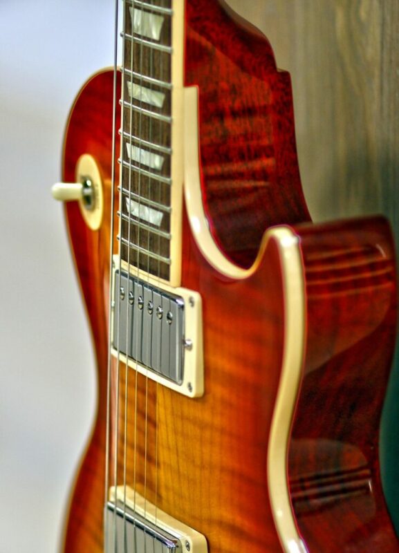 Close-up of an electric guitar with a glossy sunburst finish, cream binding, silver pickups, and strings, showing the body and part of the neck against a blurred background.