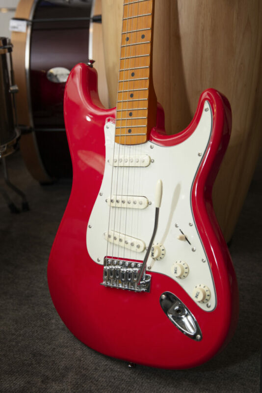 A red electric guitar with a white pickguard and maple neck rests upright on a gray carpet, with drum instruments visible in the blurred background.