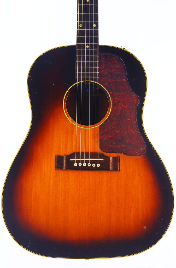 Front View Of A Gibson J-45 1957 Acoustic Guitar With A Sunburst Finish, Brown Pickguard, Six Strings, And Wooden Bridge Against A White Background.