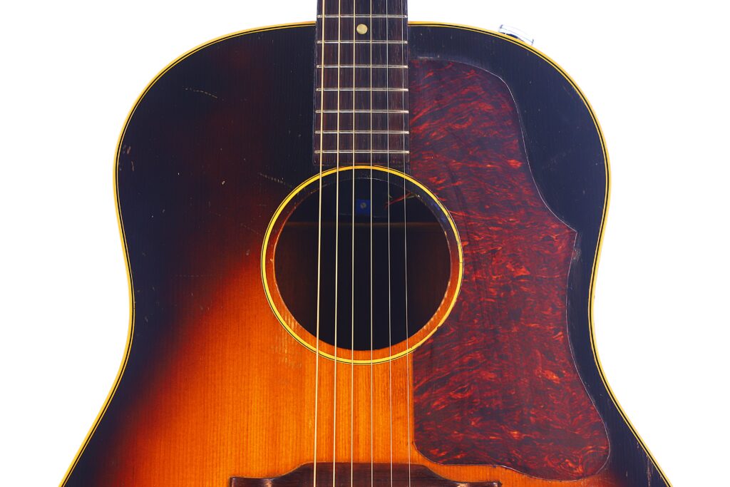 Close-Up Of A Gibson J-45 1957 Acoustic Guitar Body With A Sunburst Finish, Six Strings, A Dark Tortoiseshell-Patterned Pickguard, And Yellow-Edged Trim Set Against A White Background.